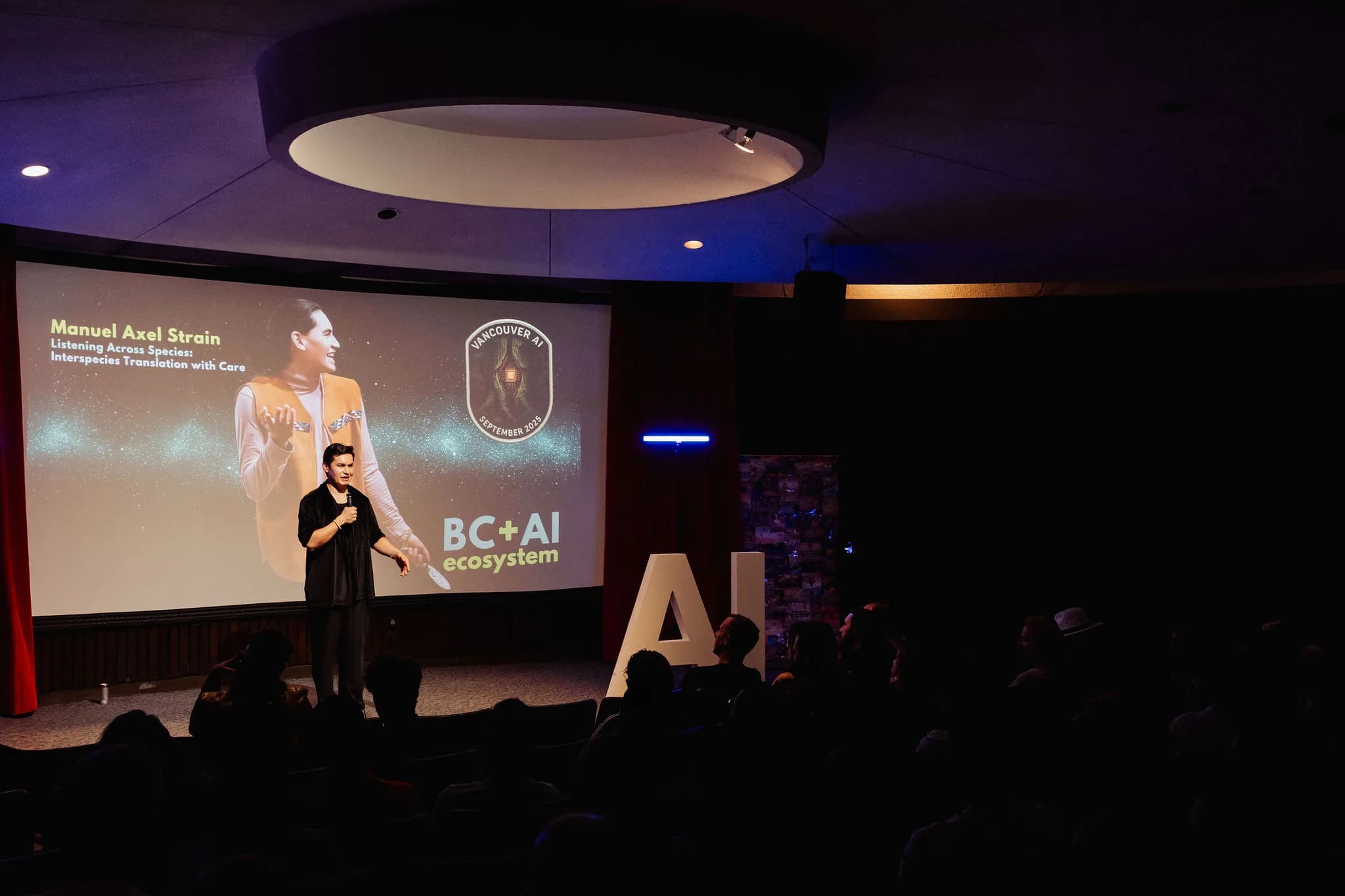 Manuel Axel Strain on stage beneath their Listening Across Species title slide at the H.R. MacMillan Space Centre. Photo by Michelle Diamond.
