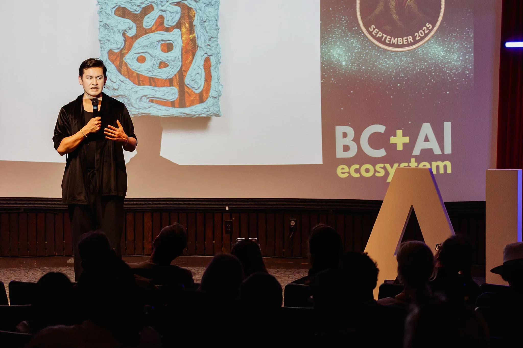 Manuel Axel Strain speaking into a microphone in front of their artwork at the September 2025 Vancouver AI meetup. Photo by Michelle Diamond.