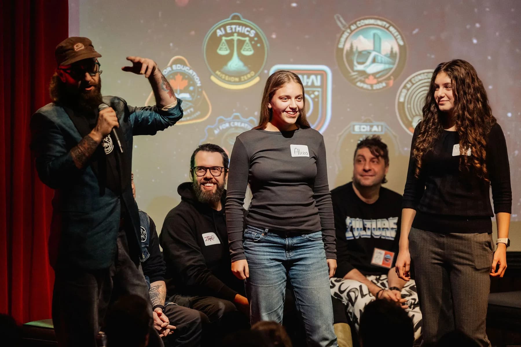 A community stage moment with badge and mission-patch designs lighting up the screen behind five people