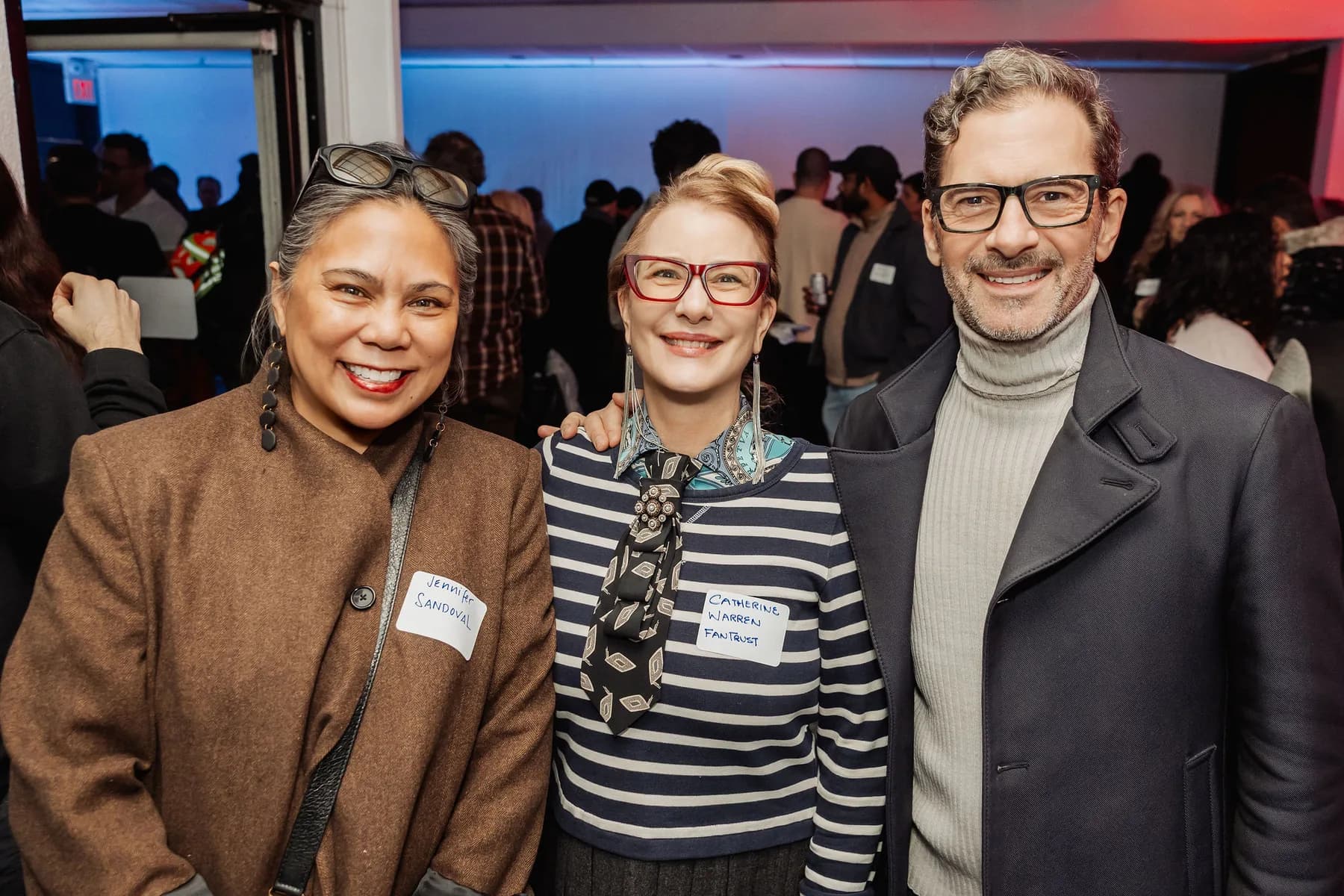 Three attendees smiling together at a packed BC + AI reception, a crowd mingling behind them