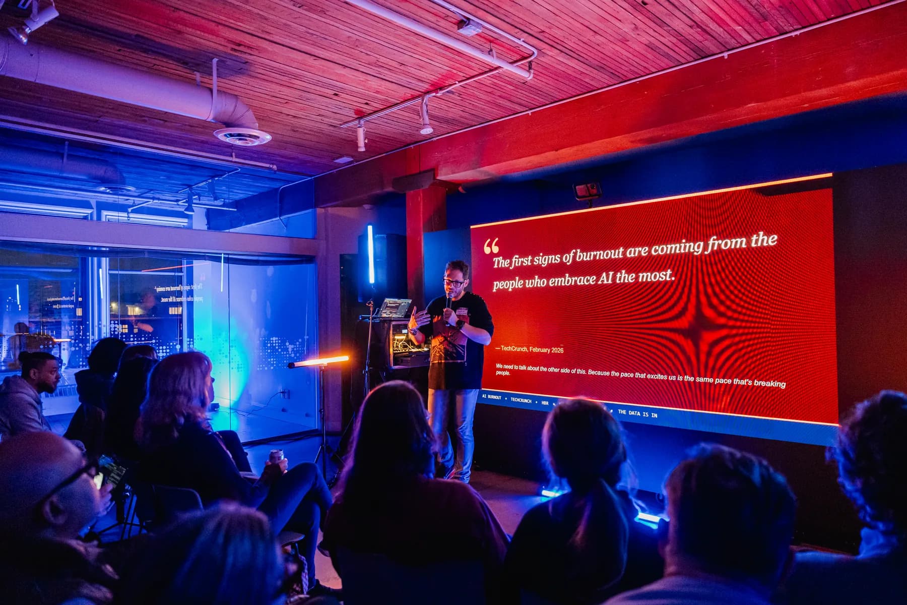 A speaker under red neon light in front of a screen reading a quote about AI burnout