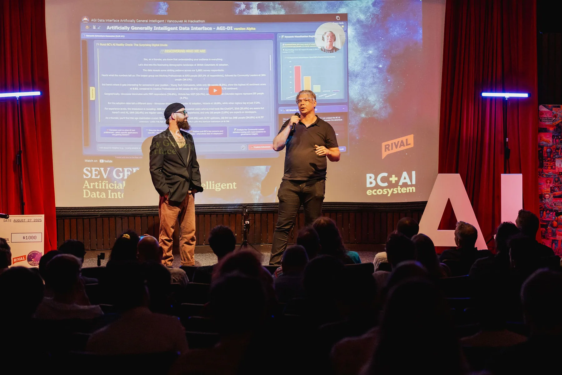 Two hackathon presenters stand on stage beside a live interface projected behind them.