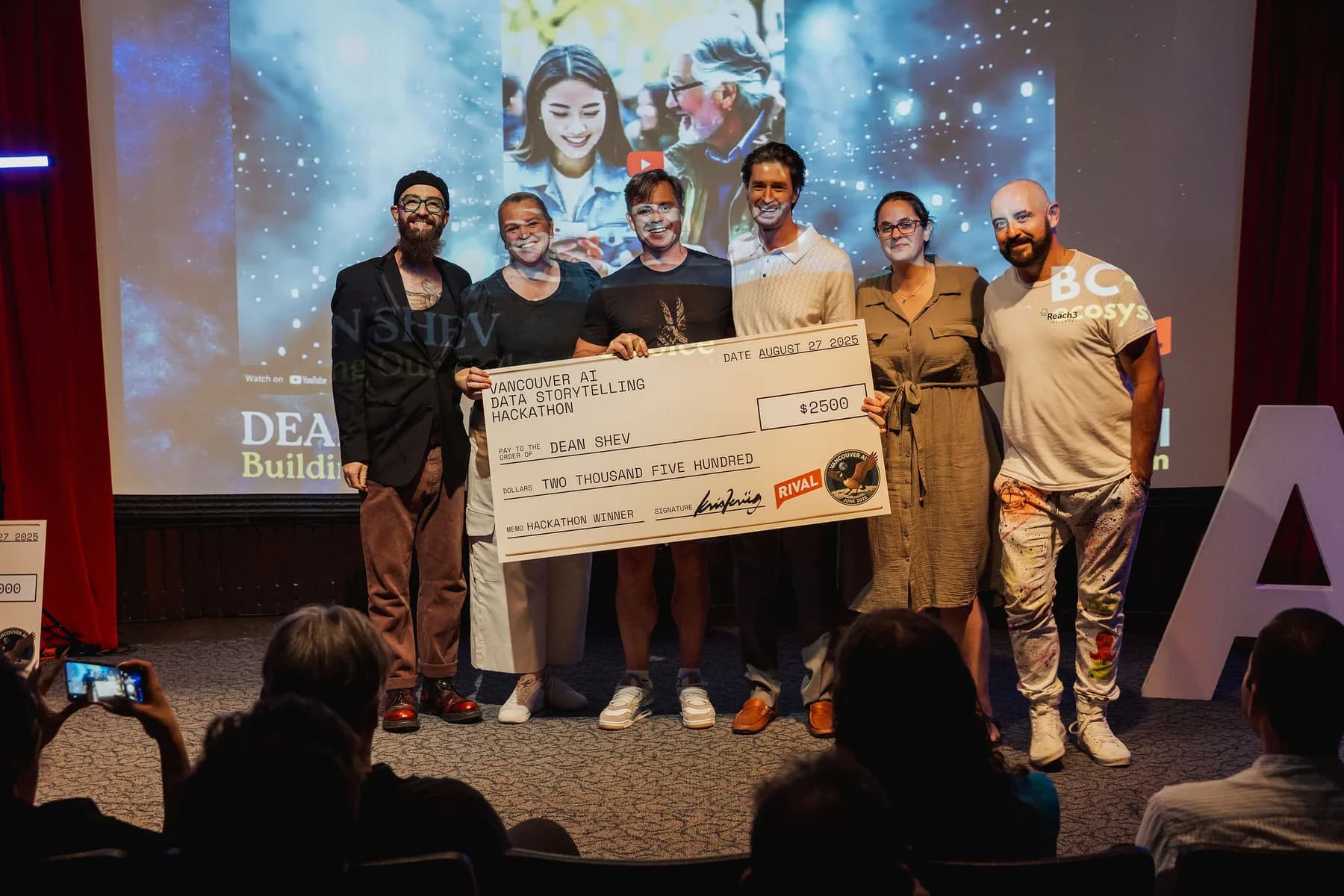 Dean Shev and the Round 3 hackathon team hold a 2,500 dollar winner cheque on stage.